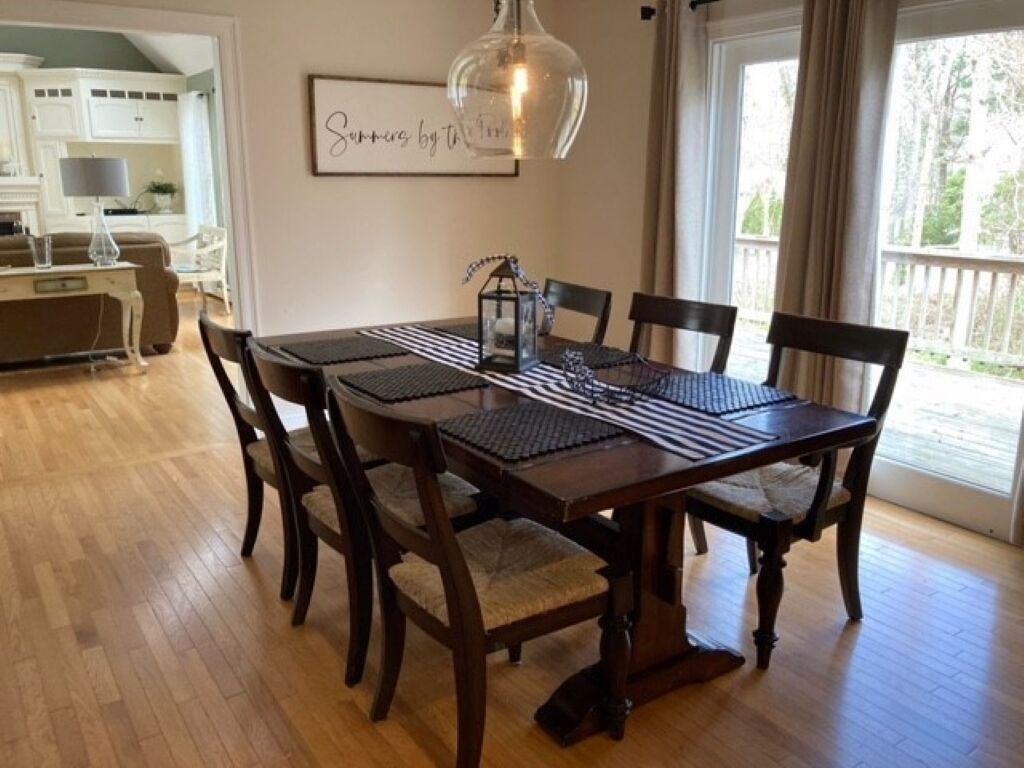 A dining area with a wooden table, six chairs, a hanging light fixture, and a framed quote on the wall.