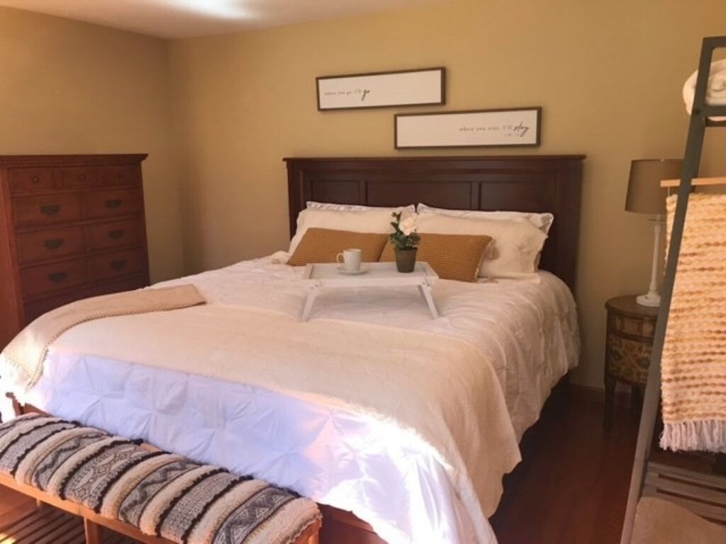 A neatly made bed with a white comforter and pillows, a bedside table with a lamp, and a wooden bench.