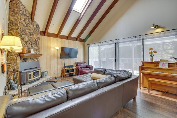 A living room with a stone wall, wooden beams, a leather sofa, a TV, a fireplace, a piano, and a large window with sheer curtains. - Ski, Hike & Fish: Big Bear Lake Home w/ Fire Pit (Big Bear Lake)