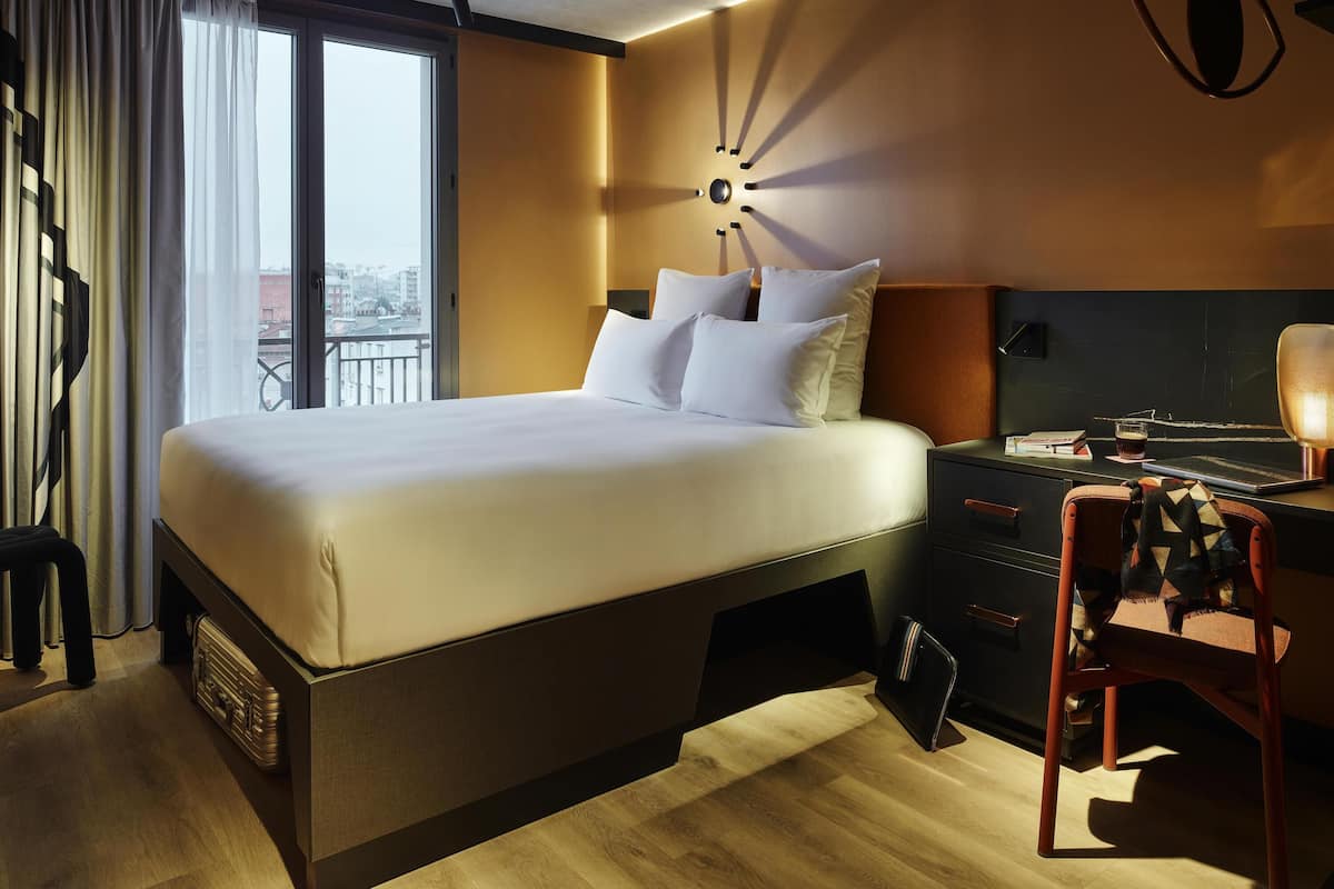 A modern hotel room with a large bed, a desk with a lamp, and a view of the city through the window.