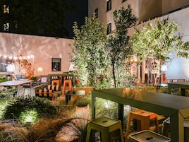 Un área de estar al aire libre con mesas y sillas, rodeada de follaje e iluminada con luces colgantes.