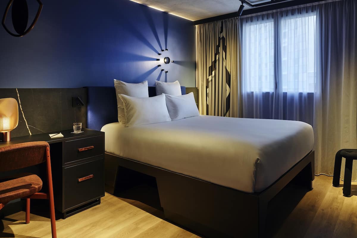 A modern hotel room with a large bed, a desk with a lamp, and a view of the city through sheer curtains.