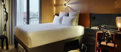 A modern hotel room with a large bed, a desk with a lamp, and a view of the city through the window.