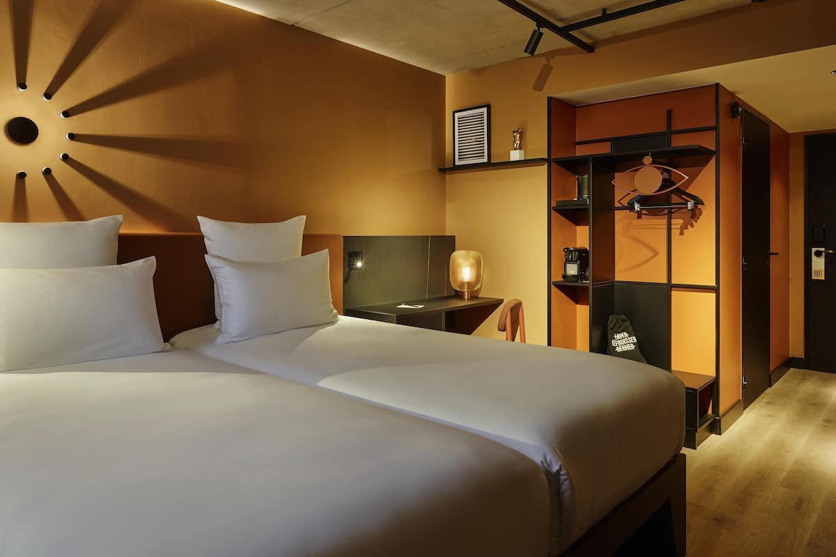 A hotel room with two beds, a nightstand, a lamp, a wardrobe, and a wall-mounted shelf.