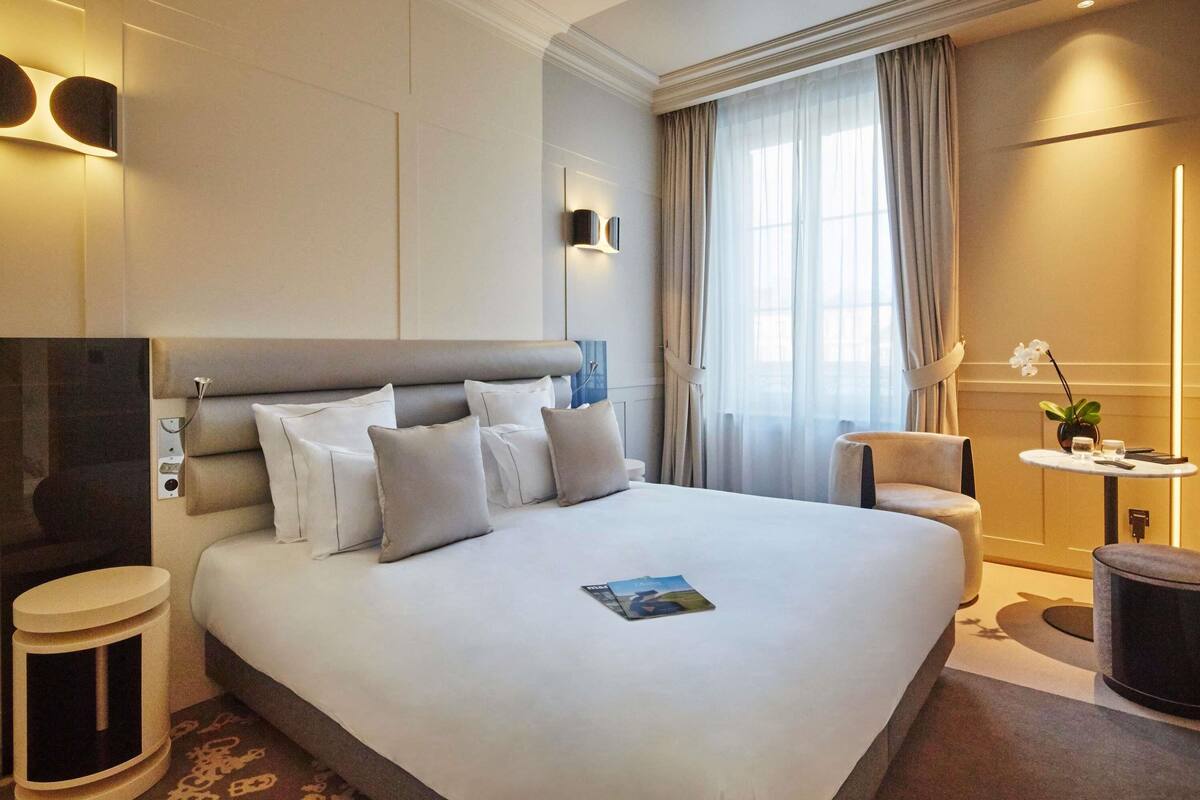 Une chambre d’hôtel moderne avec un grand lit, des tables de chevet, une petite table ronde agrémentée d’une plante, et une vue sur l’extérieur par une fenêtre.