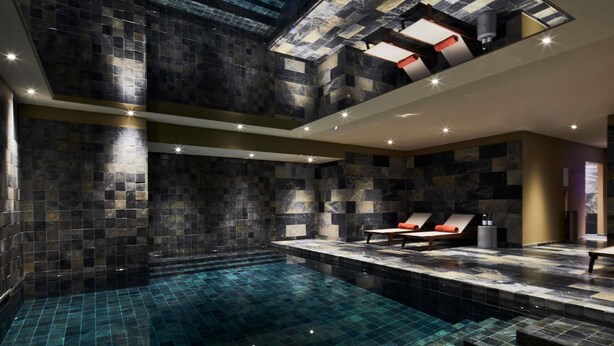 Ein moderner, überdachter Pool mit dunkel gefliesten Wänden und klarem, blauem Wasser. Liegestühle mit orangefarbenen Kissen und ein kleiner Tisch sind ebenfalls vorhanden.