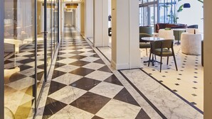 Ein Hotelkorridor mit Schachbrettboden, einer Sitzecke mit runden Tischen und Stühlen sowie Glaswänden auf der einen Seite.