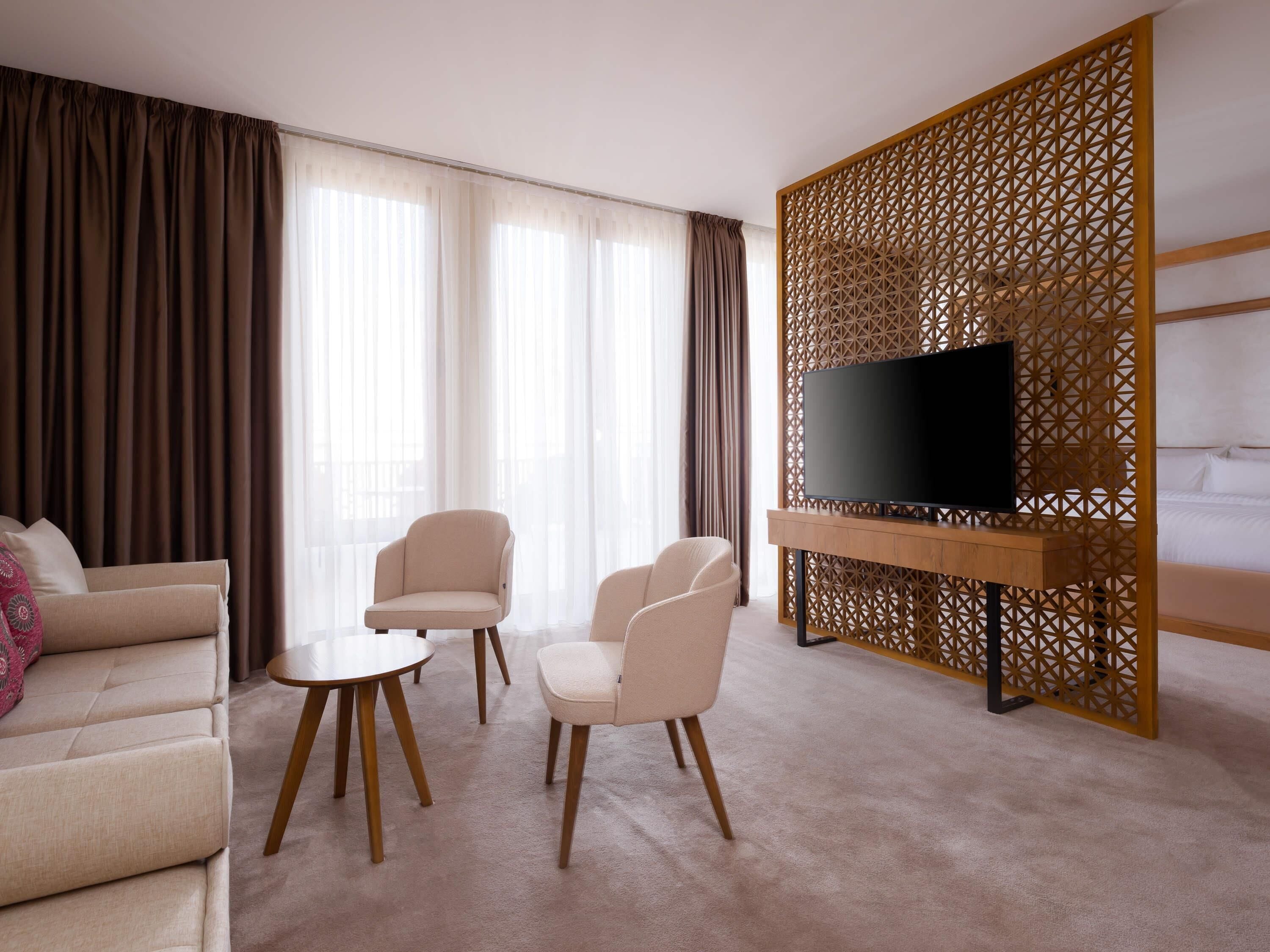 Habitación de hotel con sofá, dos sillones, una mesa de centro, televisor sobre un soporte de madera y un separador decorativo.