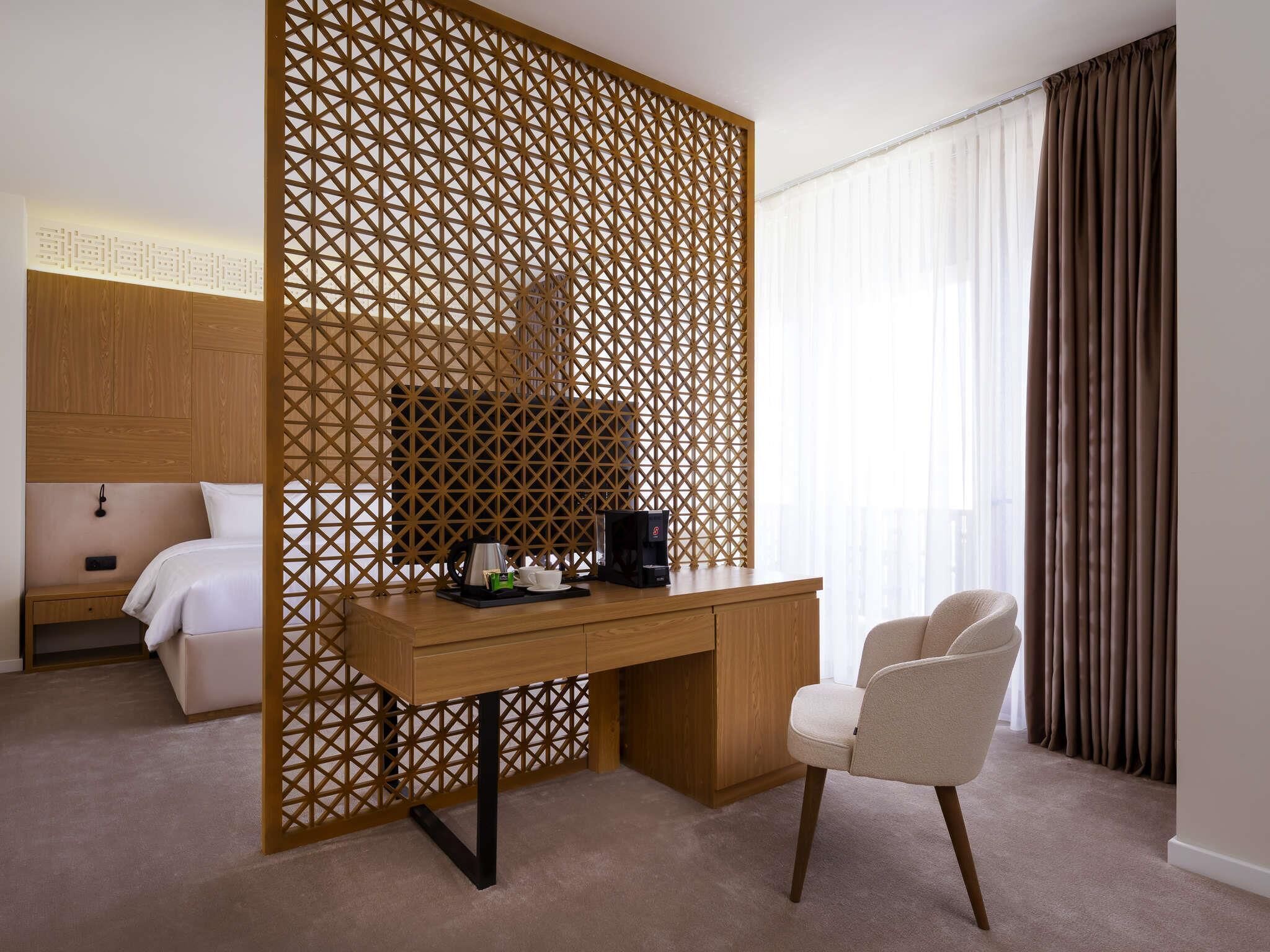 Een hotelkamer met een houten bureau, een stoel, een bed, een nachtkastje en een scheidingswand met een geometrisch patroon.