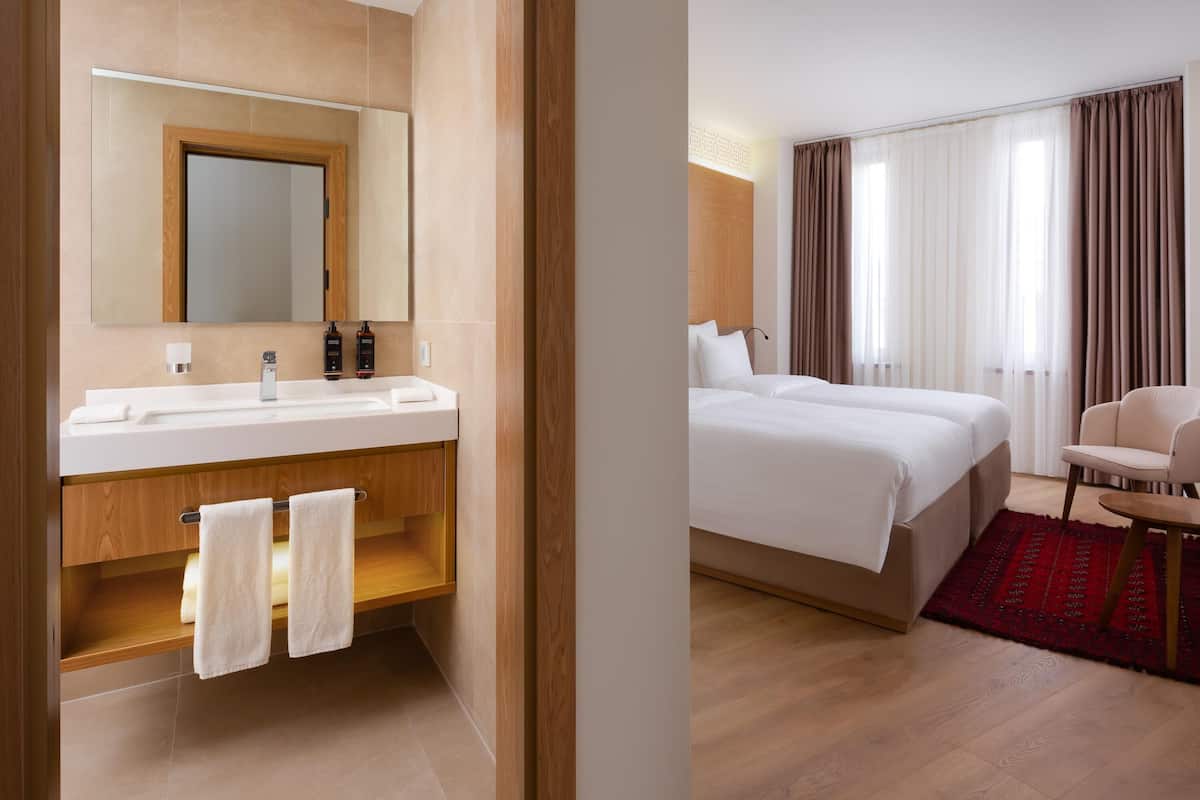 Una habitación de hotel con un tocador de madera, un espejo grande, una cama con ropa blanca, una alfombra roja y una silla.