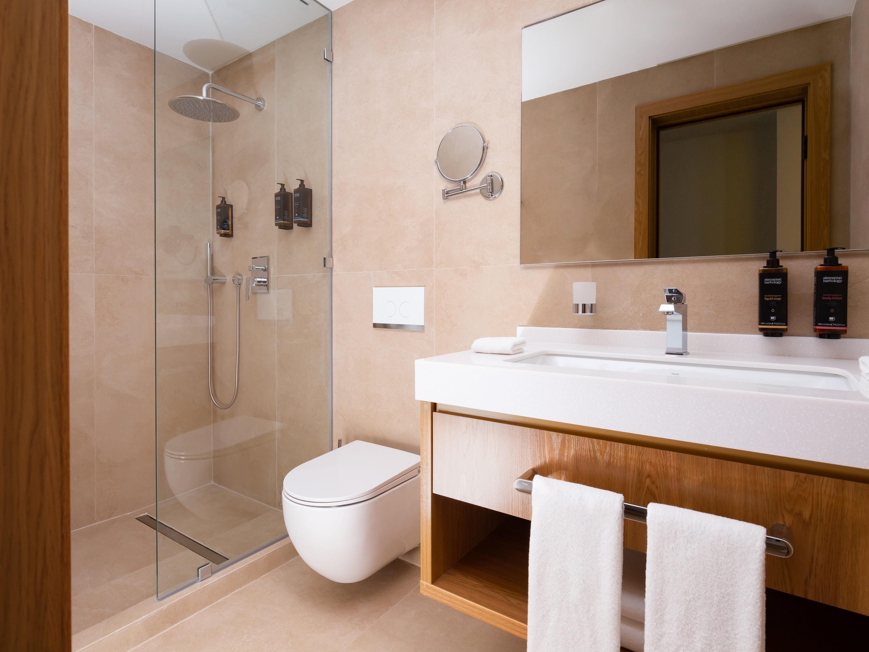 A modern bathroom with a glass-enclosed shower, a white toilet, a wooden vanity with a mirror, and two towels hanging on a rack.