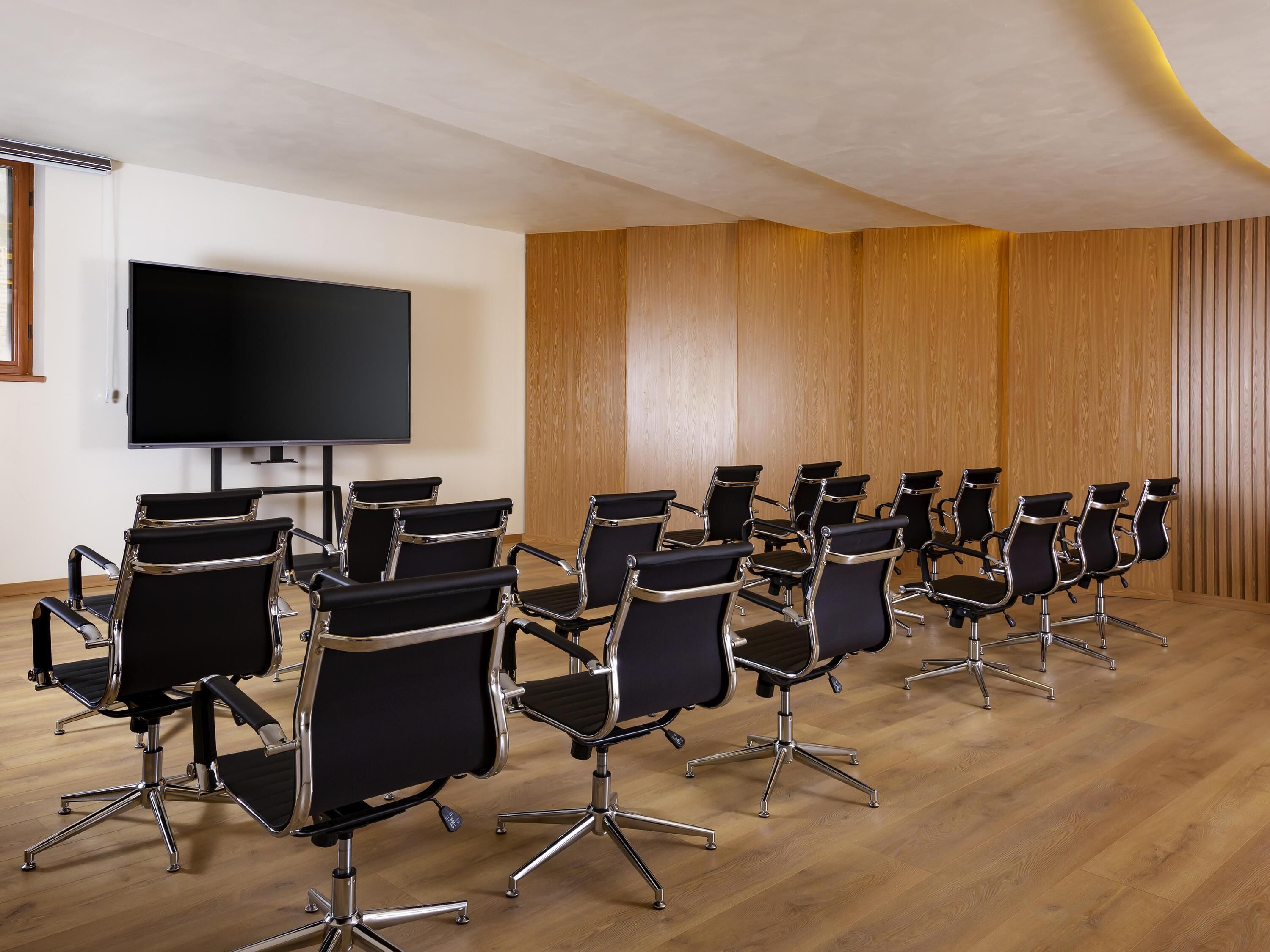 Una sala de conferencias con filas de sillas de oficina negras, un televisor de pantalla plana grande y paneles de madera en las paredes.
