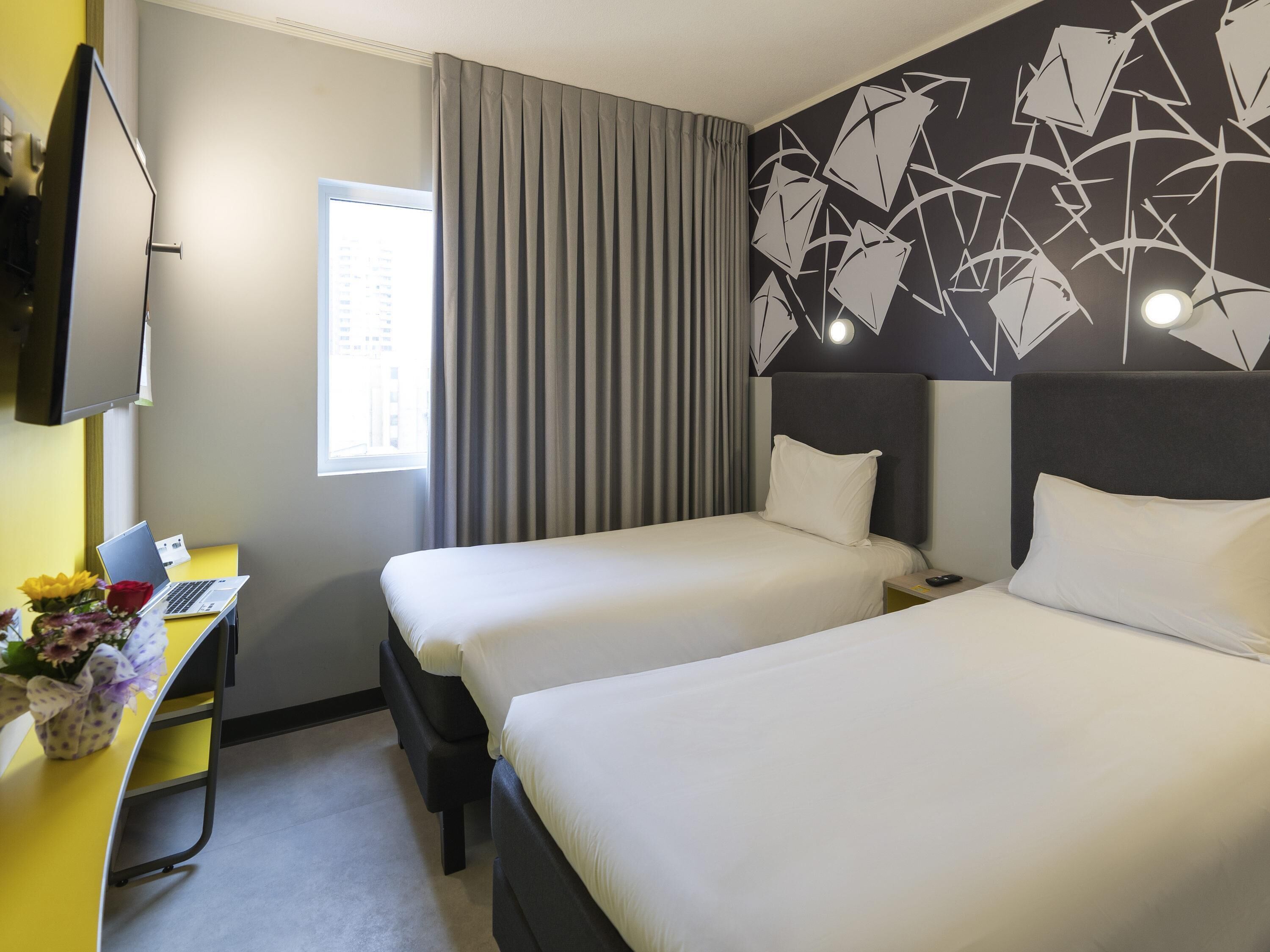 Une chambre d’hôtel avec deux lits, un bureau avec un ordinateur portable, une télévision et une fenêtre avec des rideaux.