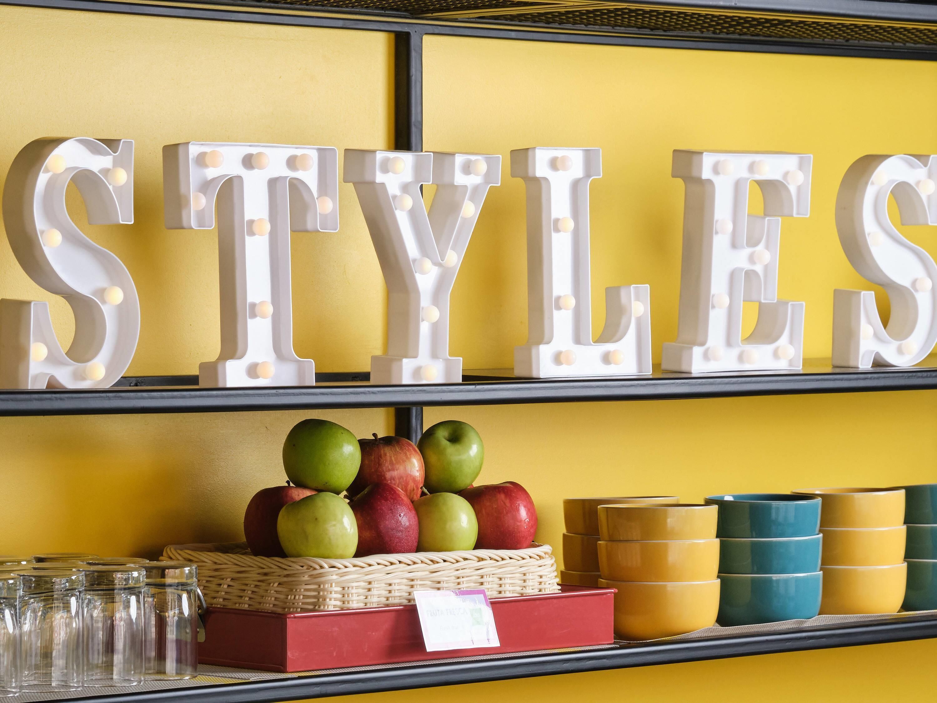 A shelf with decorative letters spelling 'STYLES,' a wicker basket with apples, and stacked ceramic cups.