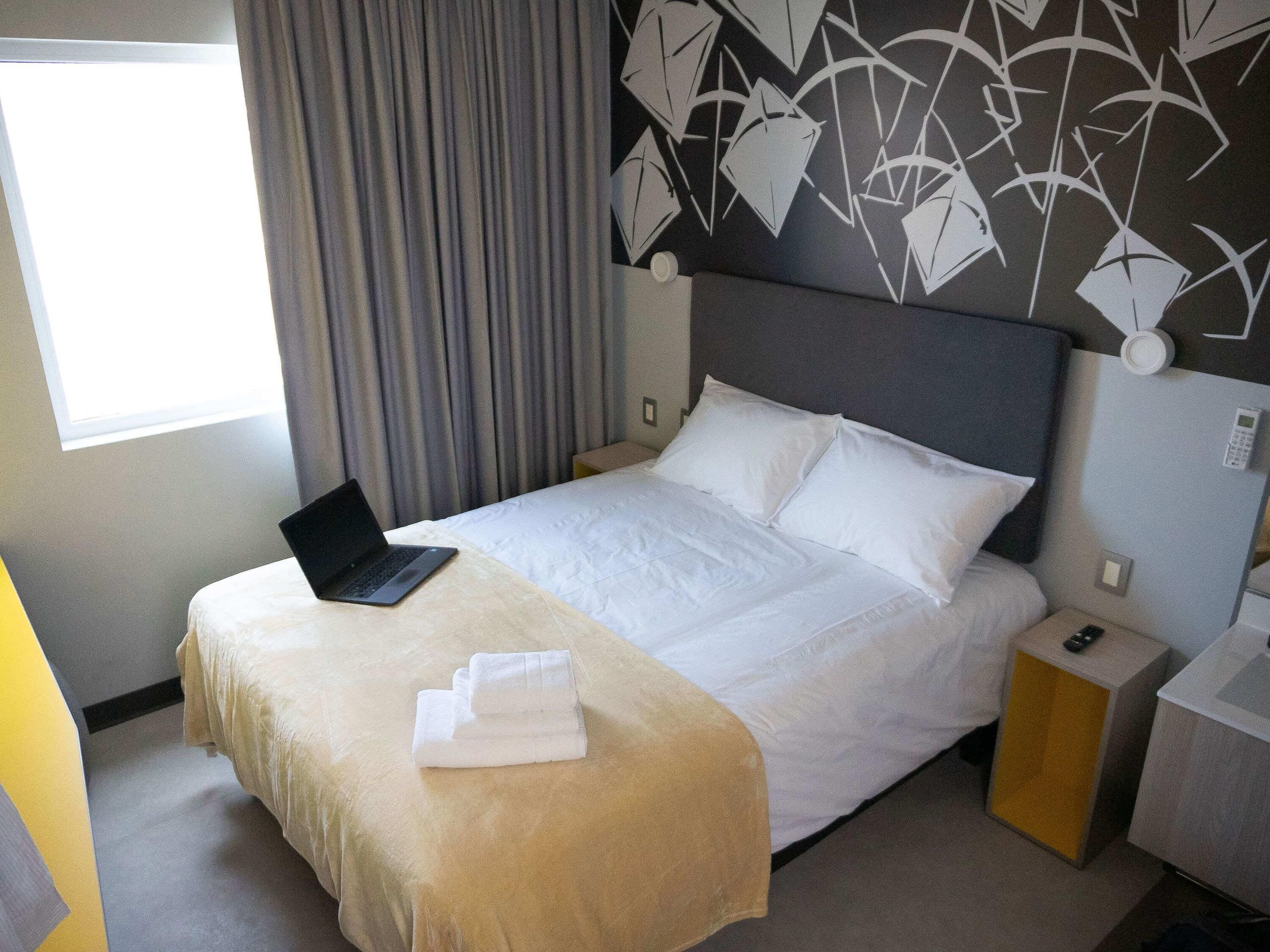 Ein Hotelzimmer mit Bett, einem Laptop auf dem Bett, einem Nachttisch mit Telefon und einem Fenster mit Vorhängen.