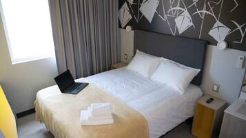 Hotelkamer met een bed, een laptop op het bed, een nachtkastje met een telefoon en een raam met gordijnen.