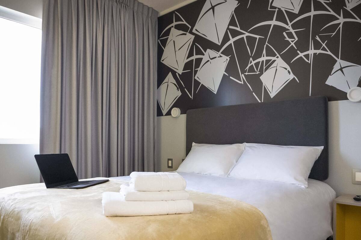 Una habitación de hotel con una cama, una laptop sobre la cama y una pared con arte abstracto.
