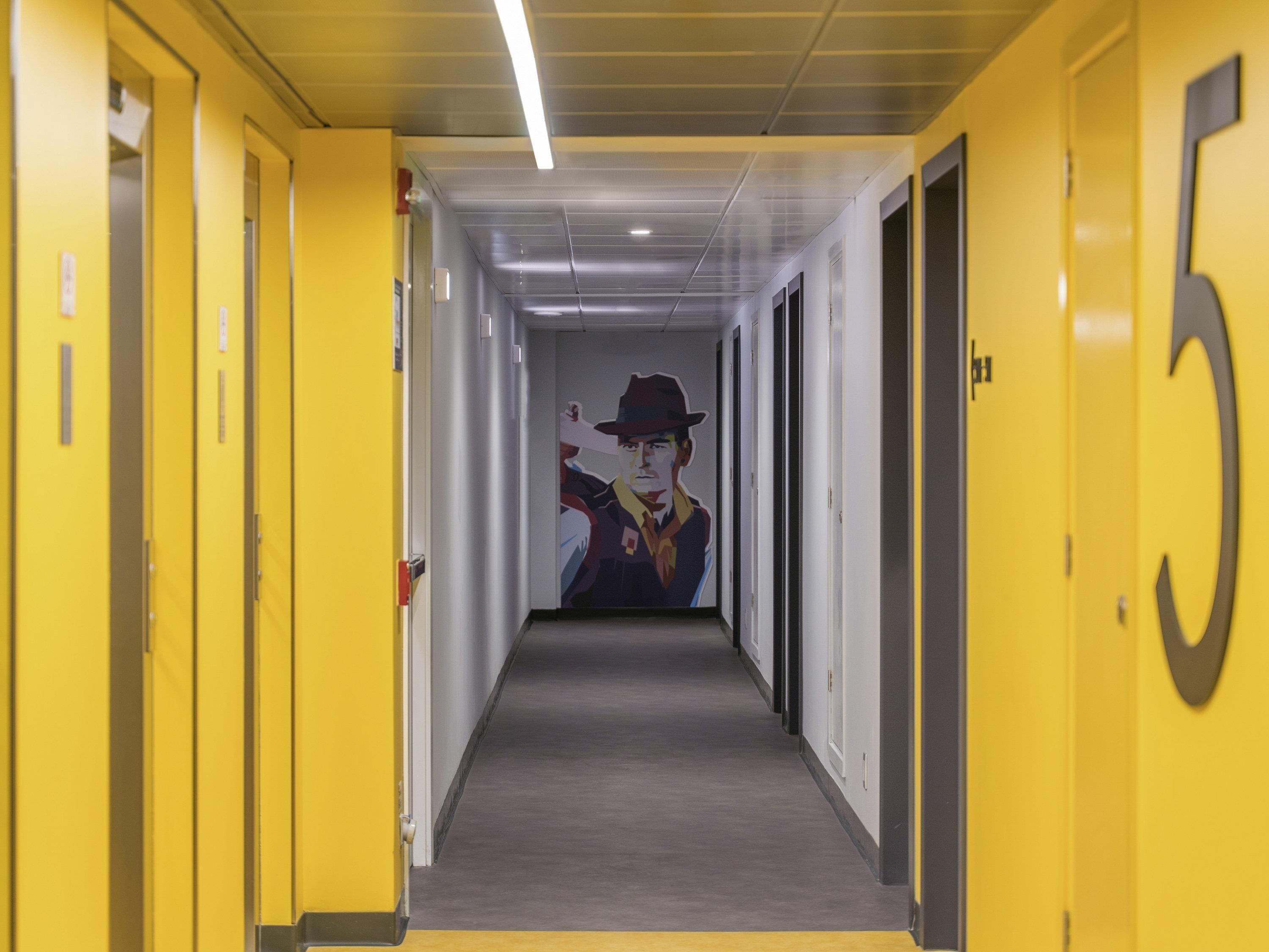 Un pasillo con puertas amarillas, un letrero con el número 5 y un mural de un hombre con sombrero.