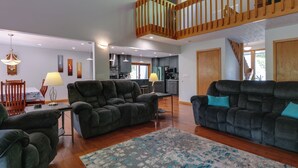 Un amplio espacio habitable con un conjunto de sofá de color oscuro, una escalera de madera y una cocina al fondo.