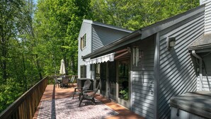 Una terraza de madera con mobiliario exterior, un espacio de patio cubierto y una casa de fachada gris.