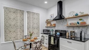 A modern kitchen with white cabinetry, a black stove, and a microwave. There are two windows with patterned curtains, a dining table with chairs, and shelves with decorative items. - Forth House – Stylish & Cozy 1 BR Apt in Springfield (Jacksonville)