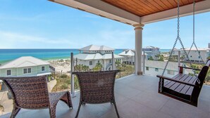 Ein Balkon mit Korbstühlen und einer Schaukel, der Blick auf Strand und Meer gewährt.