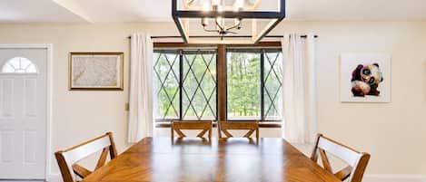 A dining room with a large wooden table, chairs, a chandelier, and a window with a view of trees.