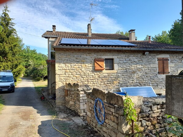 Une maison en pierre avec un toit en tuiles, une petite fenêtre avec des volets et un chauffe-eau solaire.