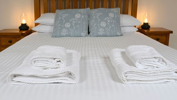 Een netjes opgemaakt bed met twee kussens, twee nachtkastjes met kaarsen, en twee opgevouwen handdoeken op het bed.