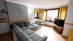 Ein Wohnzimmer mit grauem Ecksofa, einer runden Wanduhr, einem Esstisch mit Stühlen, einem Fernseher auf einem Holzboden und einem Fenster mit gelben Vorhängen.