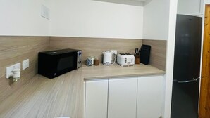 Eine kompakte Küchenzeile mit Mikrowelle, Toaster und Kaffeemaschine auf der Arbeitsplatte, einem Kühlschrank zur Rechten und einem kleinen Fenster über der Spüle.
