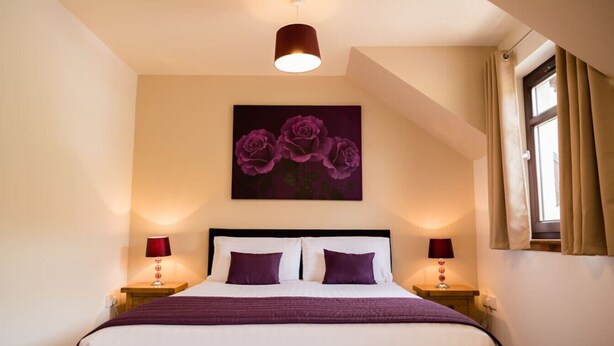 A neatly made bed with a white and purple color scheme, a painting of purple roses on the wall, and a window with curtains.