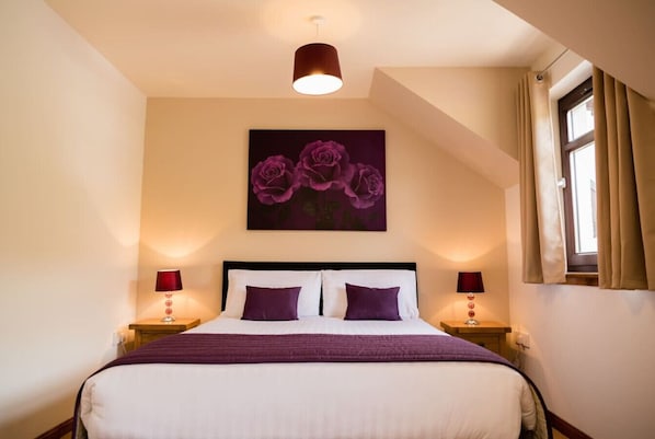 Een netjes opgemaakt bed met een wit en paars kleurenschema, een schilderij van paarse rozen aan de muur en een raam met gordijnen.