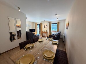 Un espace repas avec une longue table dressée pour quatre personnes, bordé de canapés et agrémenté d’un téléviseur.