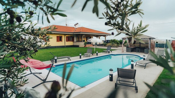 Une piscine avec des chaises longues, une maison jaune avec un toit rouge et un jardin planté d’oliviers.