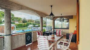 Une terrasse couverte avec une table et des chaises, un luminaire suspendu, et une vue sur une piscine et la verdure.