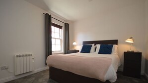 Un dormitorio con una cama grande, dos mesitas de noche con lámparas, una ventana con cortinas y un radiador.