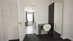 Una habitación con escritorio, silla y un espejo grande. Hay una puerta que da a otra habitación y una ventana con cortinas.