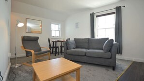 Een moderne woonkamer met een grijze bank, een houten salontafel en een bureau met stoelen. Er is een raam met gordijnen en een vloerlamp.