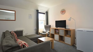 Un salon avec un canapé, un fauteuil, une télévision, une lampe et une petite table.