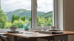 A dining table with a view of green hills through large windows. - Achintee Cottages No 1 (Highland Council)