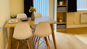 A dining area with a wooden table, four chairs, a vase with sunflowers, and a bookshelf with plants.