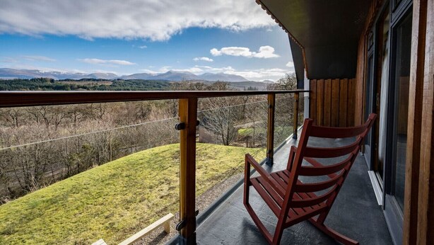 Un balcón con una silla de madera, vista a una colina cubierta de hierba y una cadena montañosa distante.