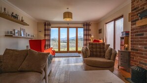 Un salón con sofá, sillón y una mesa de centro. Hay una chimenea, una estantería y vistas al exterior a través de amplios ventanales.