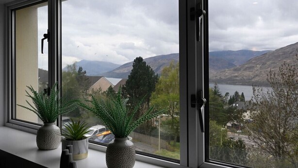 A window with a view of a lake, mountains, and houses. - Highland Lookout in Fort William (Highland Council)