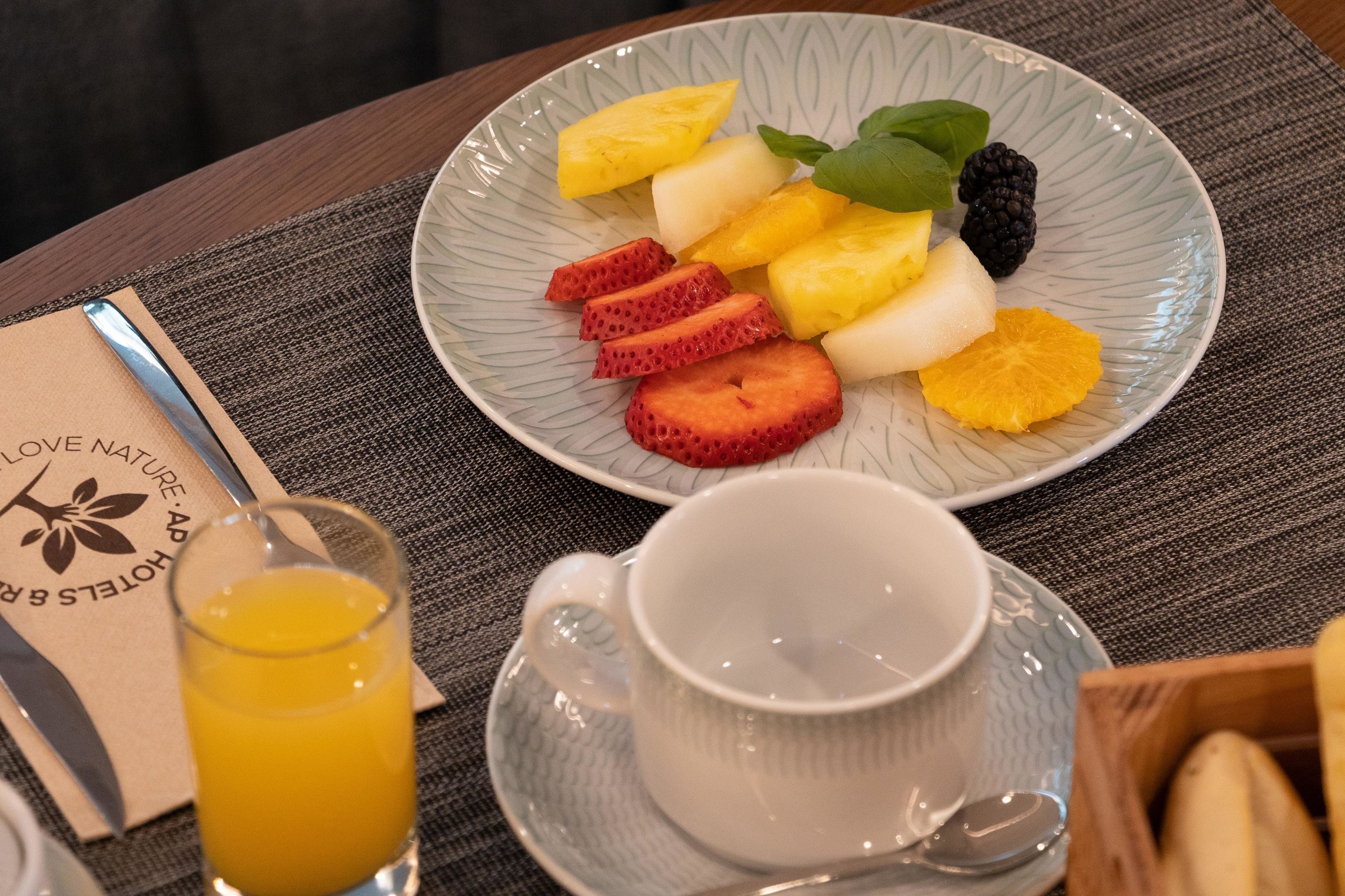 A plate of fruit including strawberries, pineapple, and blackberries, a glass of orange juice, and a cup on a table.