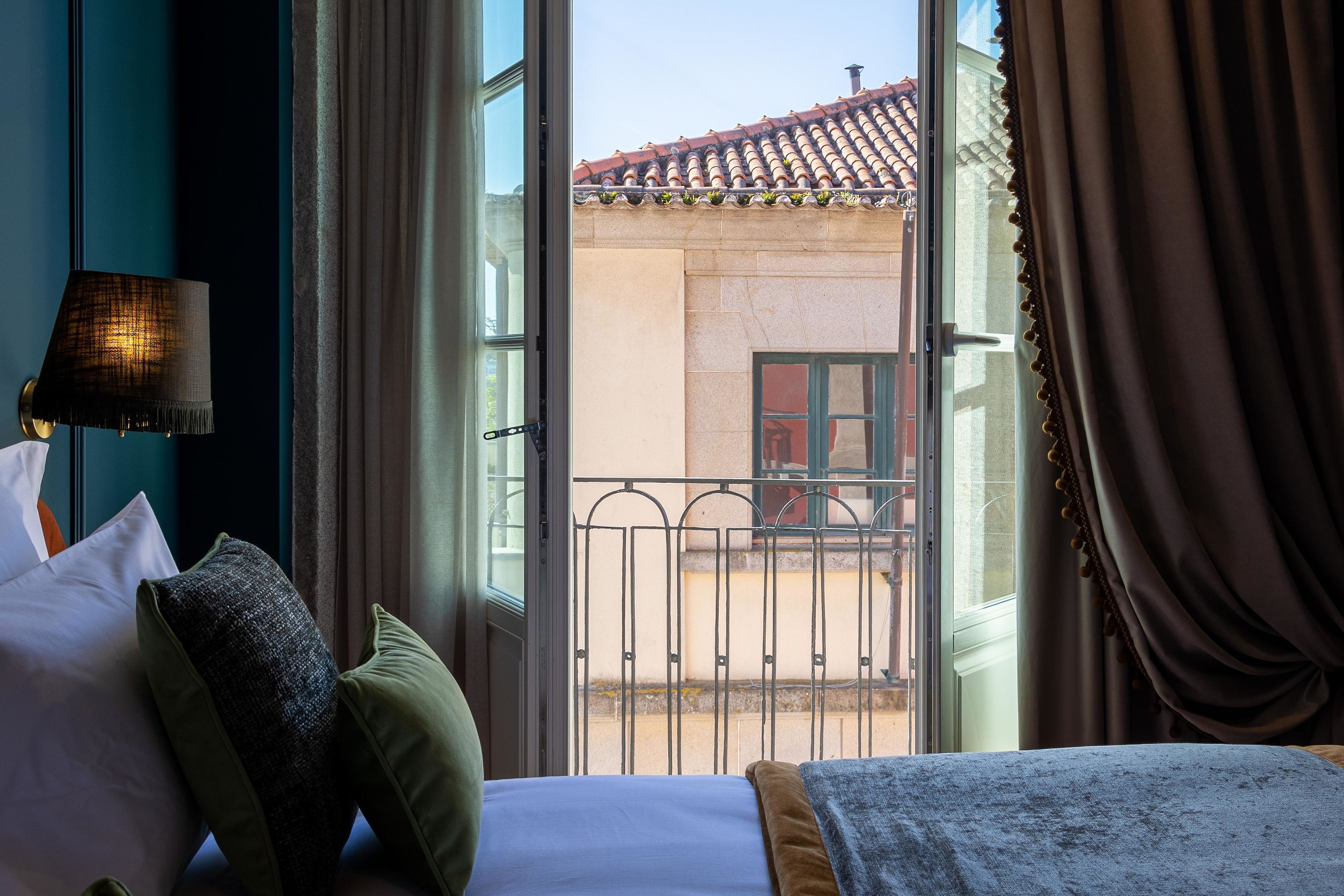 Habitación de hotel con cama, almohadas, lámpara y vista a un balcón con barandilla y un edificio al fondo.