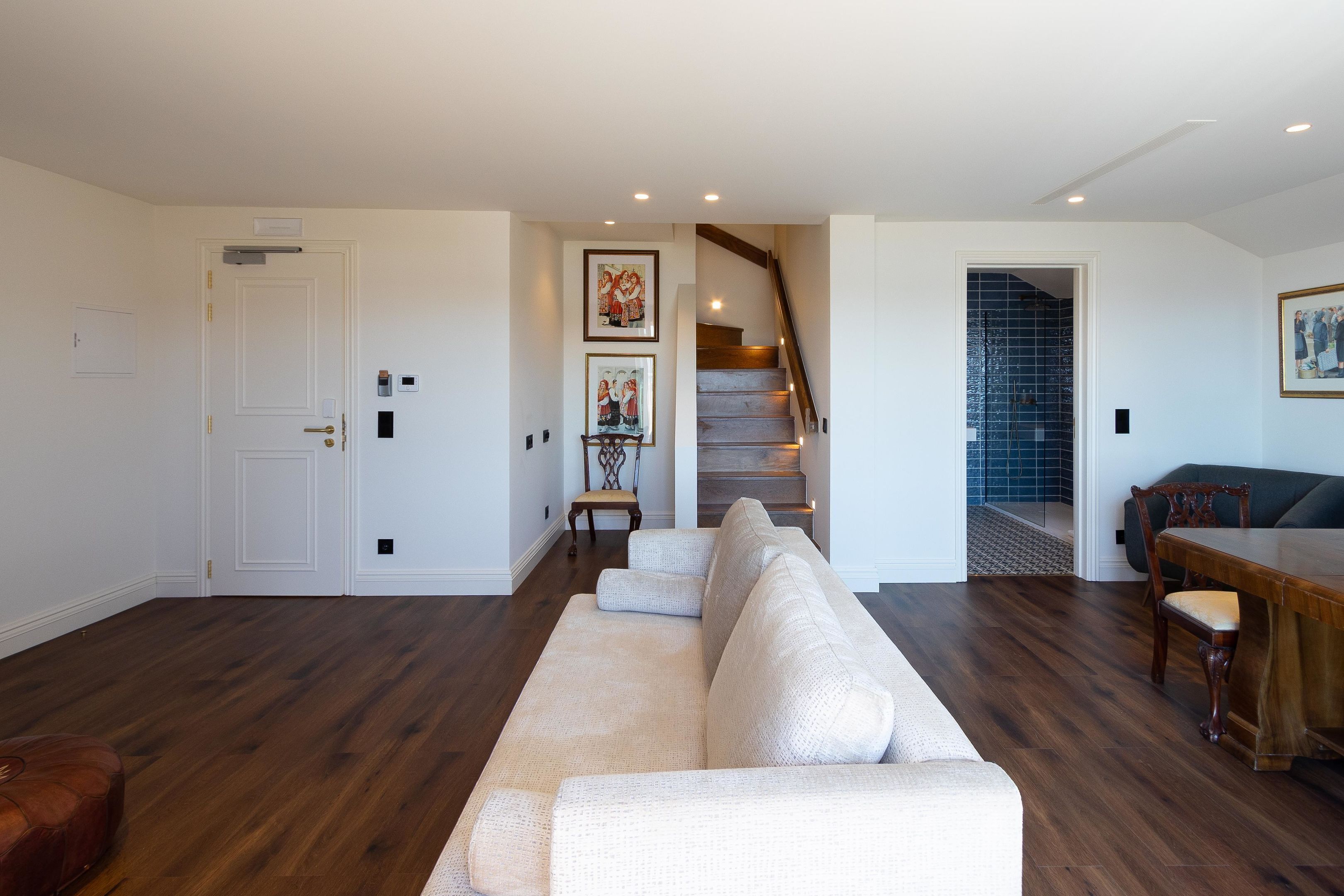 Un salon comprenant un grand canapé d’angle beige, une table à manger en bois avec des chaises, un escalier avec une rampe en bois et une porte avec une plaque nominative.