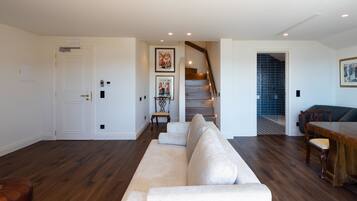Un salon comprenant un grand canapé d’angle beige, une table à manger en bois avec des chaises, un escalier avec une rampe en bois et une porte avec une plaque nominative.