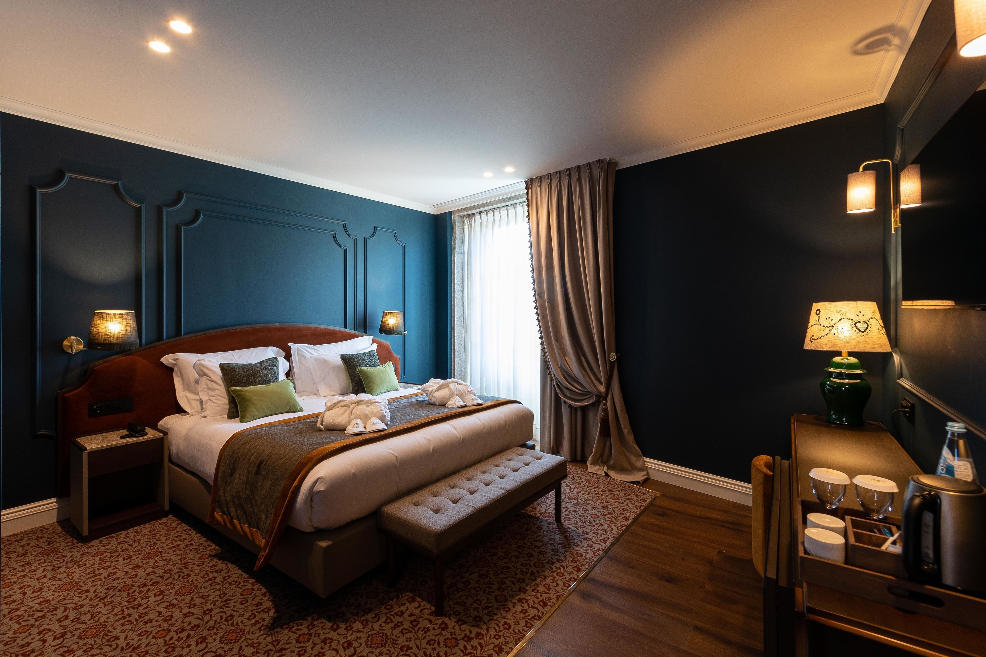 A modern hotel room with a large bed, bedside lamps, a small table with cups and a kettle, and a view of the outdoors through a window.